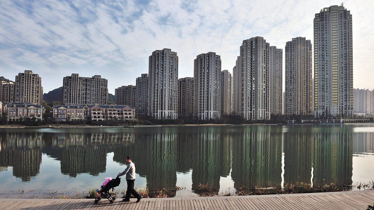 A local Chinese resident pushes a baby stroller along a lake in front of a cluster of residential apartment buildings in Changsha city.