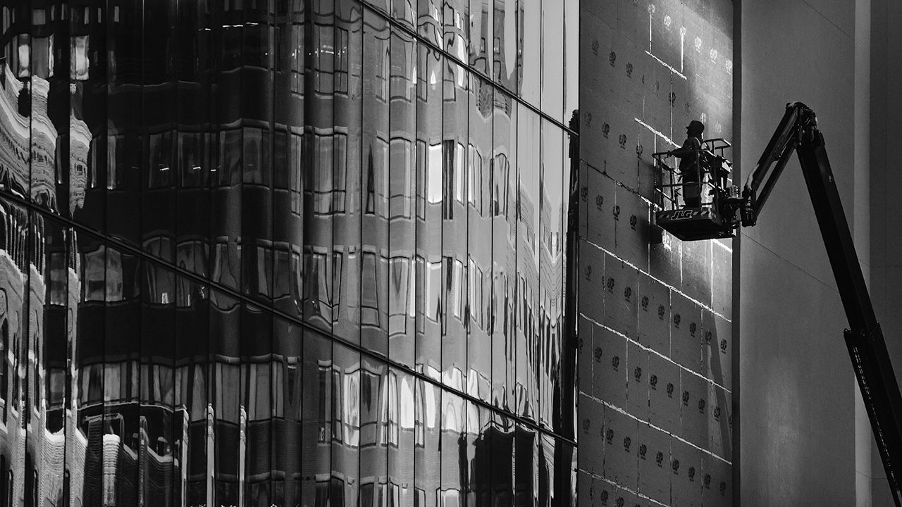 The reflections of buildings are seen in the windows of a skyscraper that a construction worker works on.