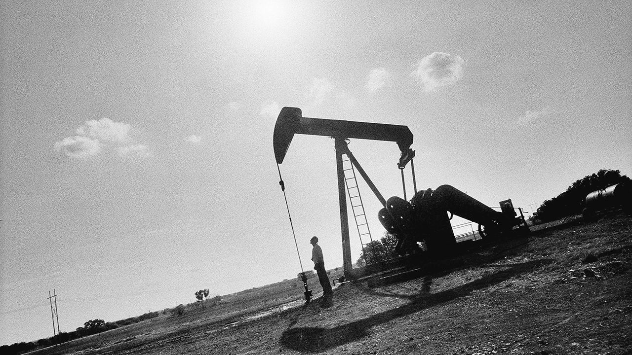 The silhoutte of a man looking up at a pump jack in an oil field.